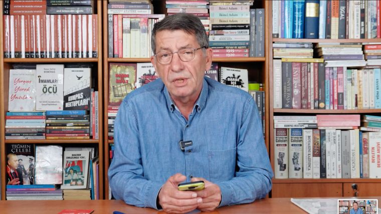 A man holds a cell phone and speaks in front of a book shelf.